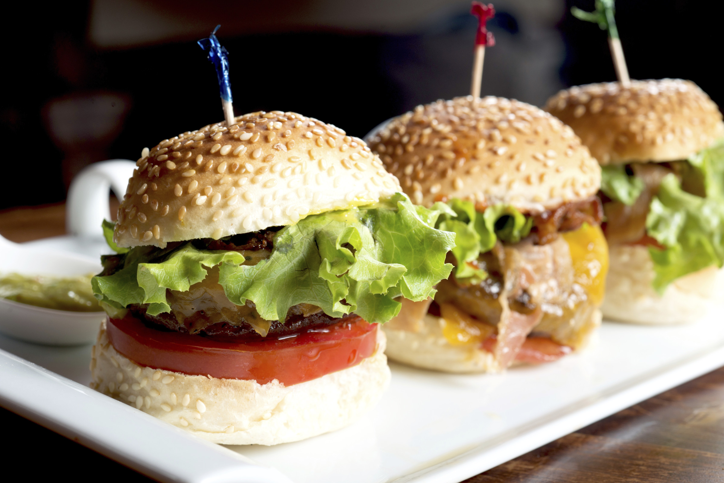 Three gourmet hamburgers with sesame seed buns, fresh lettuce, and tomato slices, served on a white platter, highlighting the culinary offerings relevant to restaurant mystery shopping evaluations.